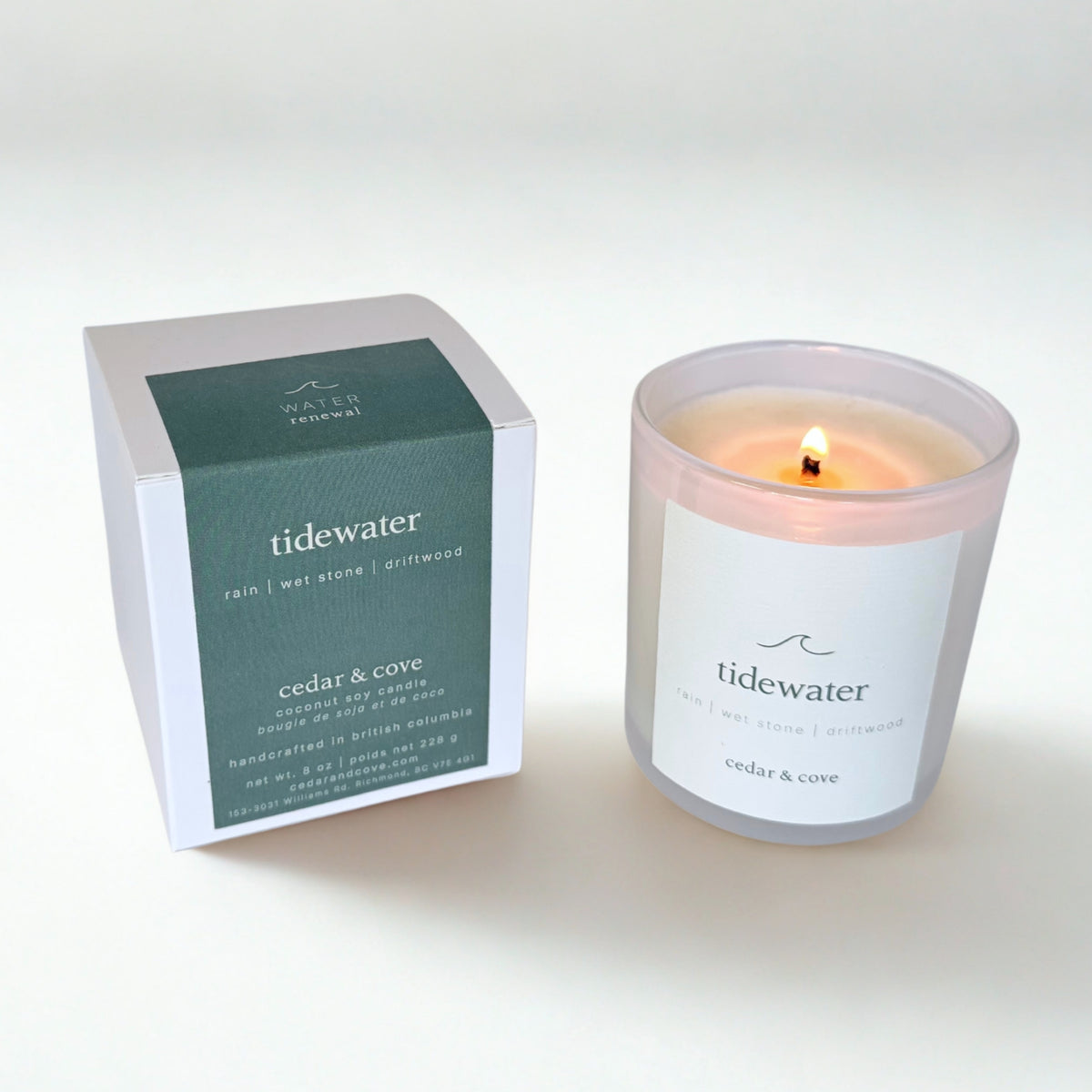 A lit 'Tidewater' candle by Cedar & Cove in a frosted glass jar next to its white and dark green packaging box on a clean white background.