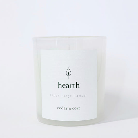 A frosted glass jar candle called Hearth by Cedar & Cove with cedar, sage, and amber scent notes. Photographed on a clean, white background.