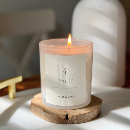 A lit 'hearth' scented candle by Cedar & Cove in a frosted glass jar, resting on a rustic wooden slice coaster. The candle features a white label and is set against a softly blurred background with warm, natural light casting shadows.