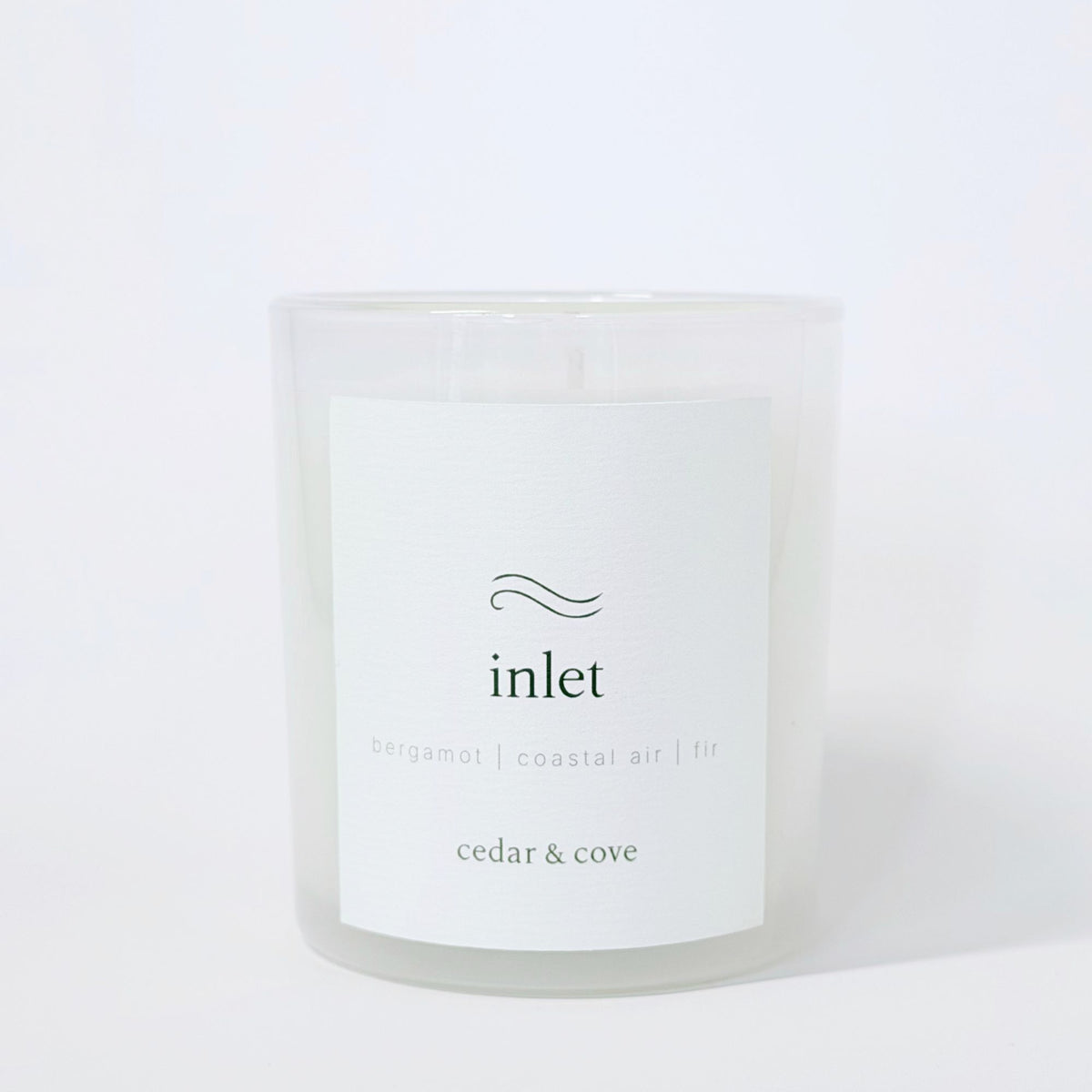A frosted glass candle called 'Inlet' by Cedar & Cove with a white label featuring bergamot, coastal air, and fir scent notes. Photographed on a clean white background.