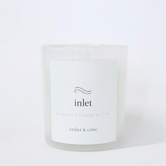 A frosted glass candle called 'Inlet' by Cedar & Cove with a white label featuring bergamot, coastal air, and fir scent notes. Photographed on a clean white background.