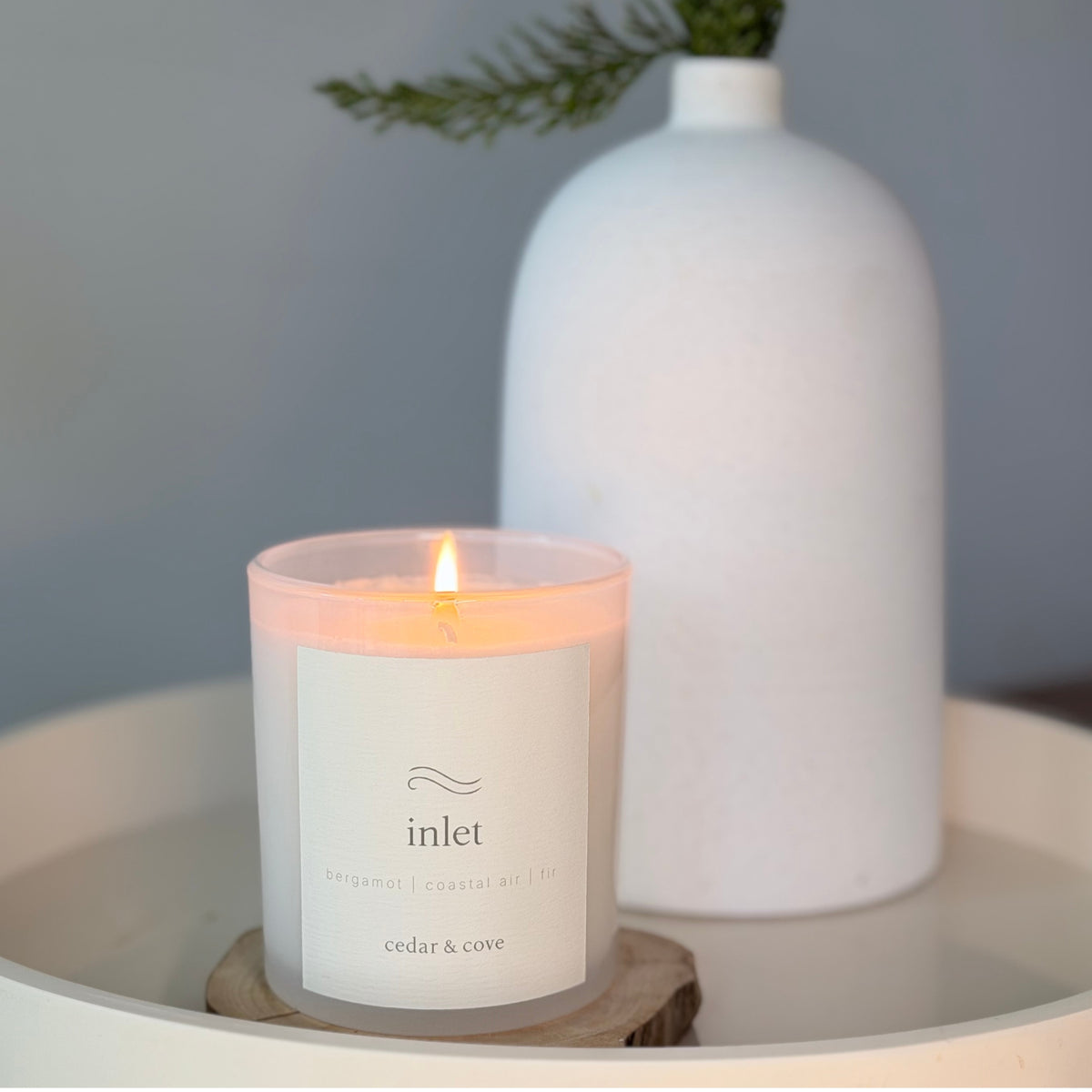 A lit Inlet candle by Cedar & Cove in a round, frosted glass vessel sits on a wooden coaster on a white tray, with a large white ceramic vase in the background against a soft gray wall.