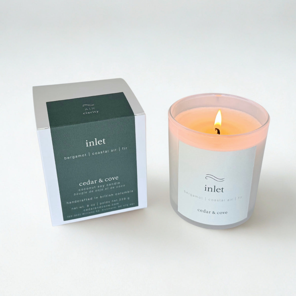 A lit 'Inlet' scented candle by Cedar & Cove in a frosted glass jar with a white label, shown next to its white and dark green packaging box.