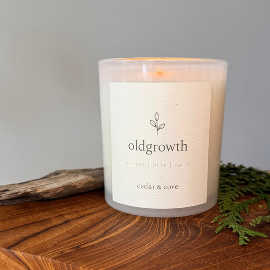 A lit scented candle by Cedar & Cove in a frosted glass jar sits on a wooden surface with a neutral gray background, a piece of driftwood, and green foliage. The label reads 'oldgrowth' with juniper, pine, and resin scent notes. 