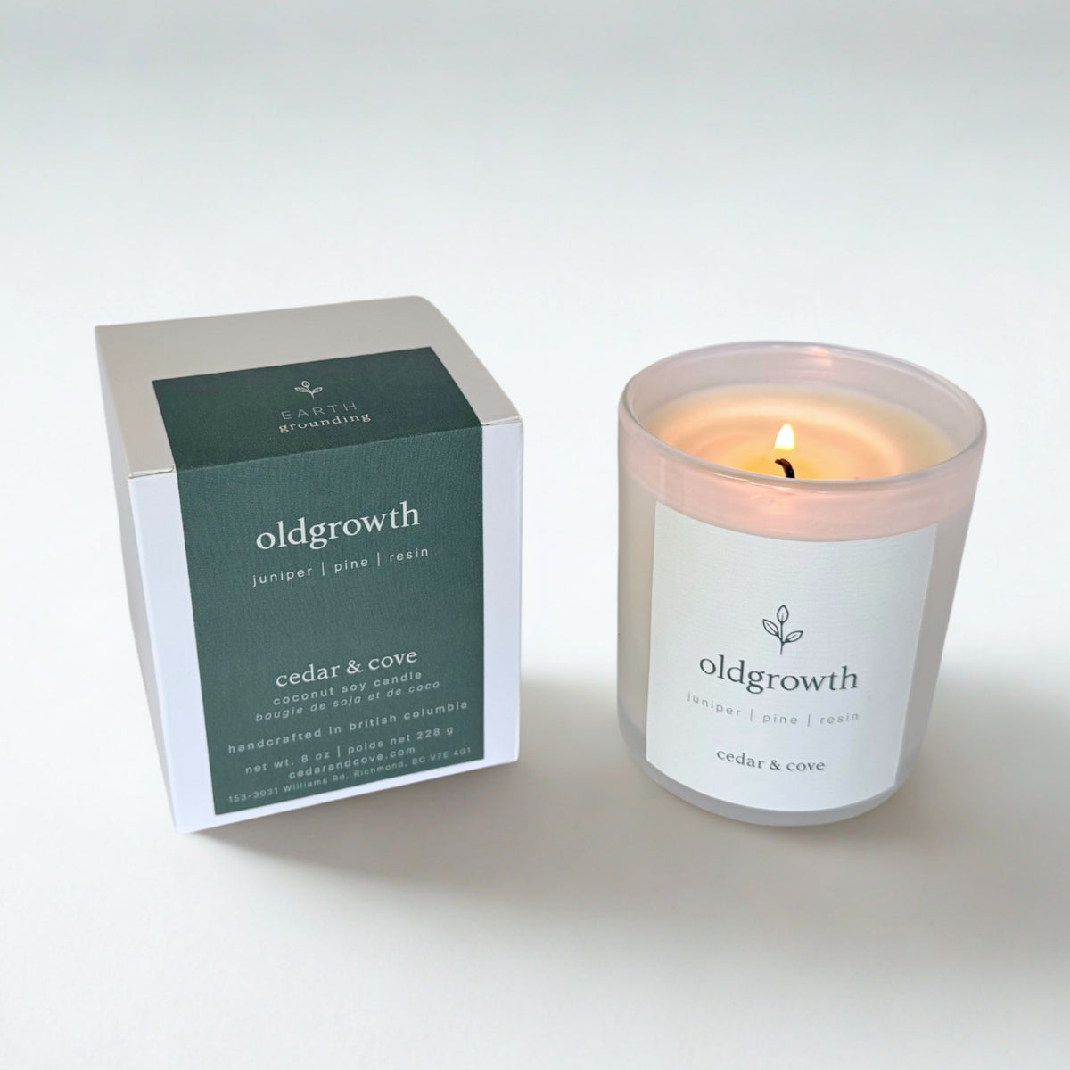 A lit candle in a frosted glass jar next to its product box. The candle and packaging feature a white label and a dark green accent panel with the scent name 'oldgrowth' and brand name 'Cedar & Cove'.