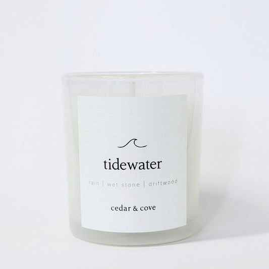 A frosted glass candle Called 'Tidewater' by Cedar & Cove with a white label featuring rain, wet stone, and driftwood scent notes. Photographed on a clean white background.