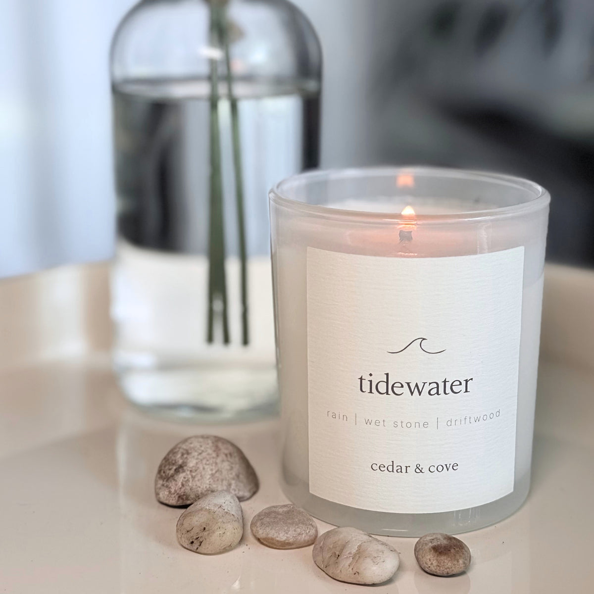 A lit 'Tidewater' candle by Cedar & Cove in a frosted glass jar with a white label, placed on a light surface with small stones in front and a blurred glass vase in the background.
